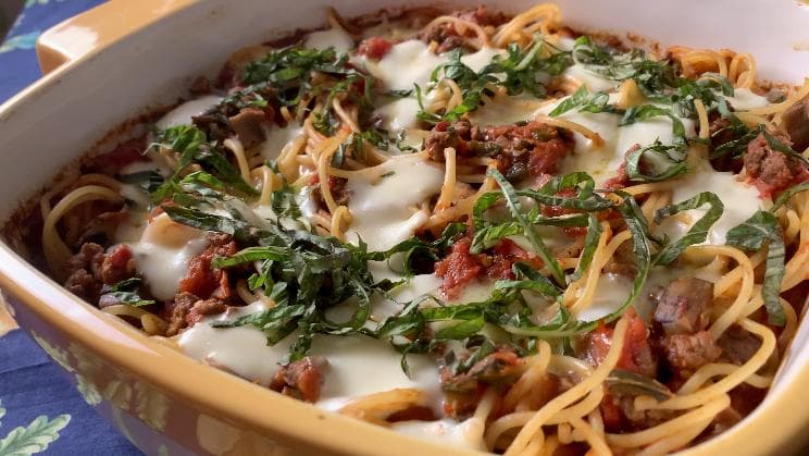 Baked spaghetti with meat sauce, melted cheese, and fresh basil in a white ceramic dish.