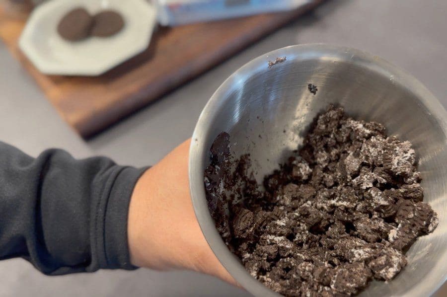 Hand holding metal bowl filled with crushed Oreo cookies for latte preparation.