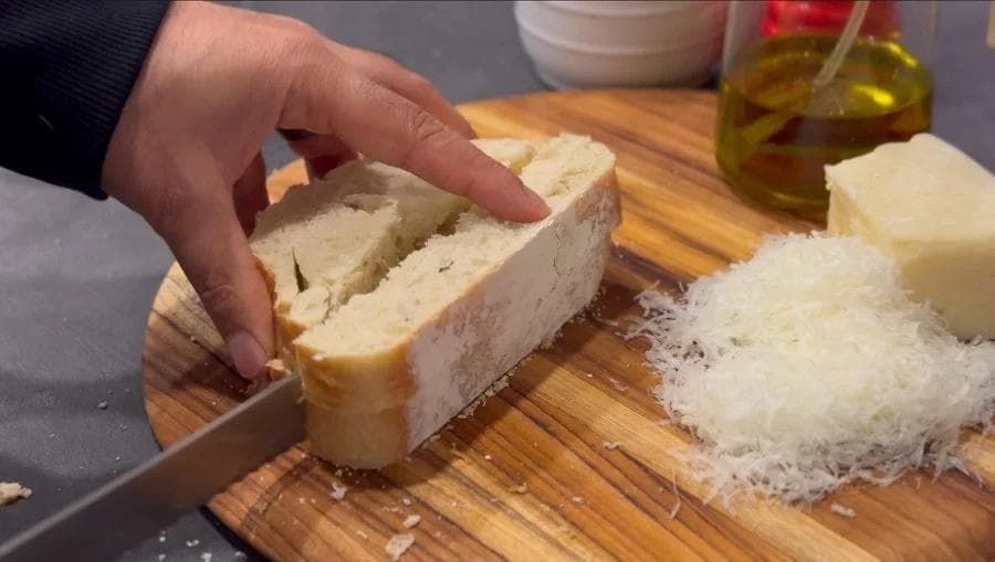 Hand slicing fresh bread on a wooden cutting board next to grated Pecorino Romano cheese and olive oil.