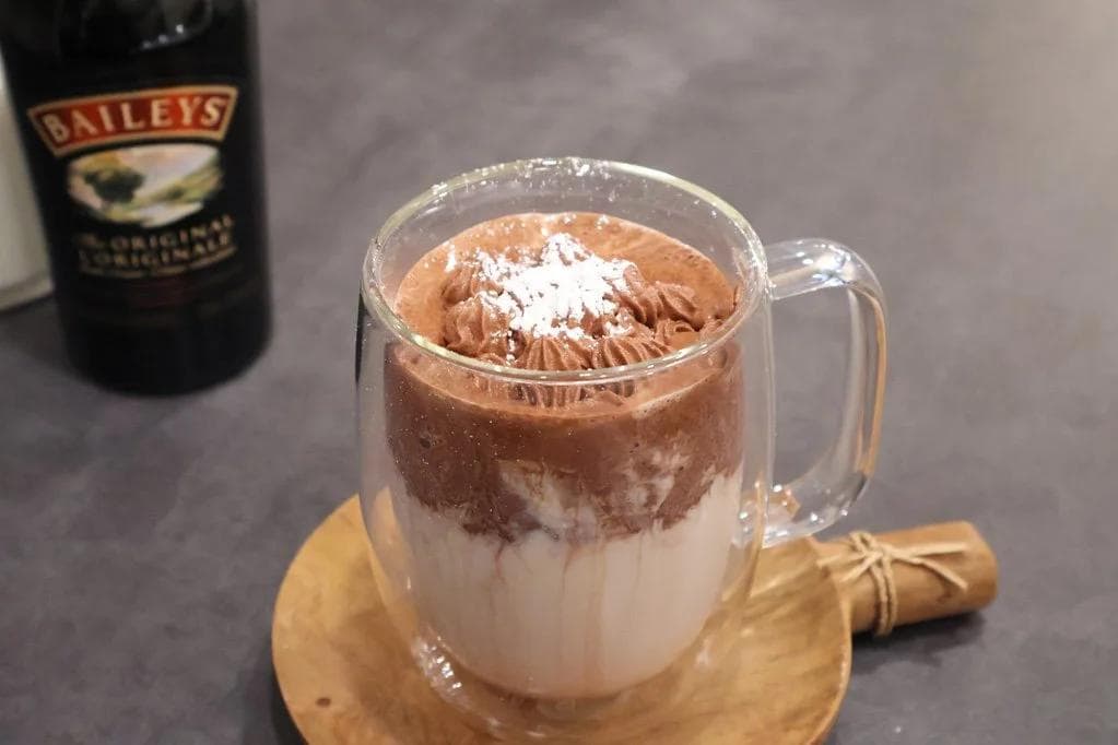 Whipped Baileys hot chocolate in a clear mug with cocoa powder and whipped cream on top, next to a Baileys bottle.