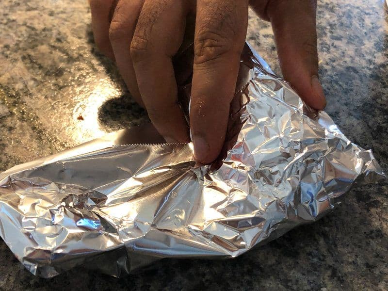 Hands wrapping a chicken kathi roll in aluminum foil on a granite surface.