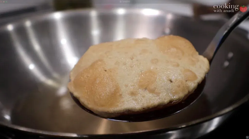 Golden puffy poori frying in oil on a slotted spoon held over a stainless steel wok.