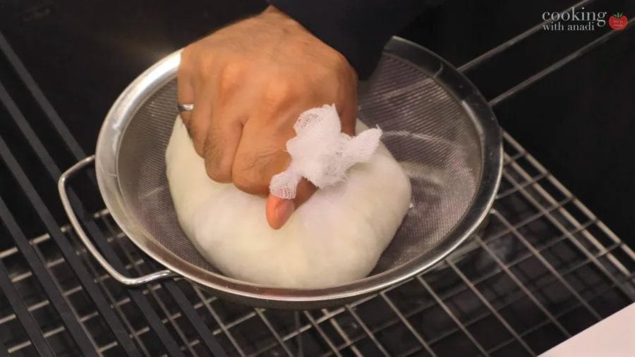 Hand squeezing paneer in cheesecloth over a colander to remove excess whey.