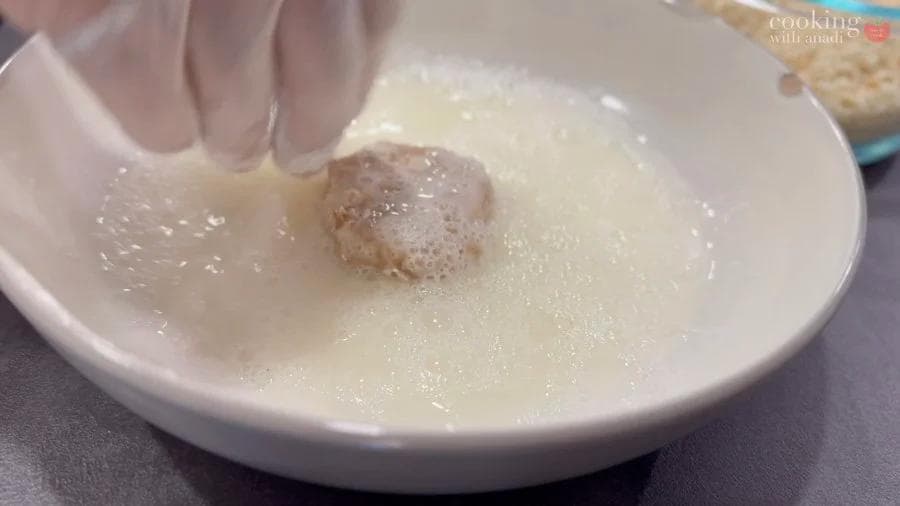 Raw chicken nugget being pressed into breadcrumbs in a white bowl during coating process.