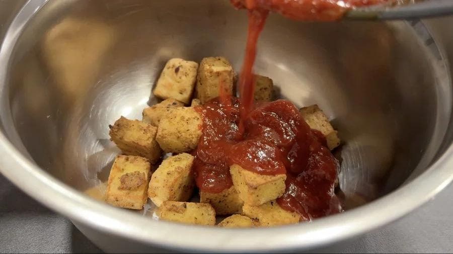 Crispy cubed tofu being coated with glossy red Korean sauce in a stainless steel bowl.