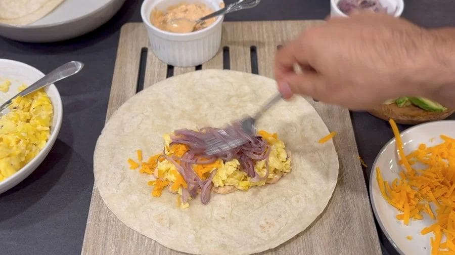 Hands folding a flour tortilla filled with scrambled eggs, shredded cheese, and red onions on a wooden board.