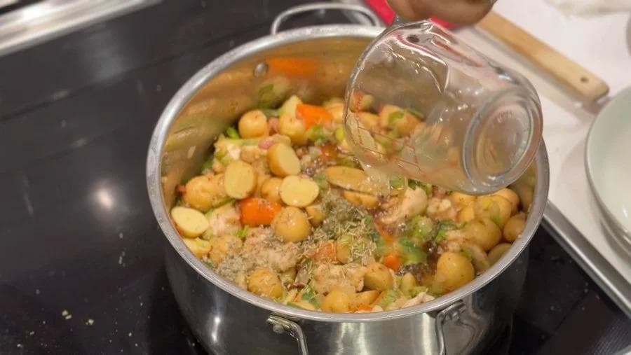 Pot of chicken stew with potatoes, chickpeas, carrots, peas, and ground meat in broth being stirred with a glass pitcher.