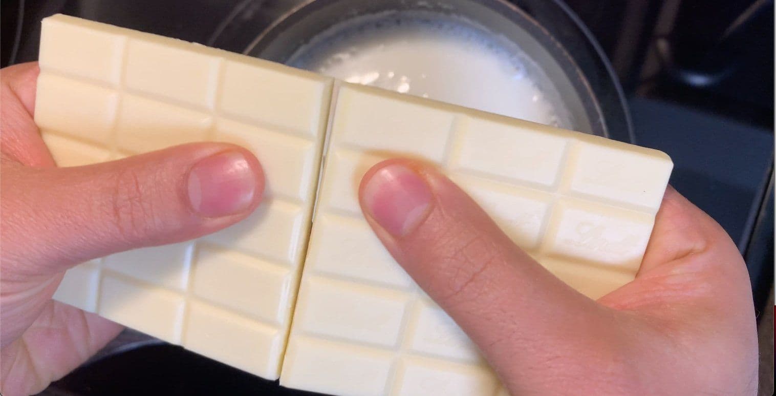 Hands breaking white chocolate bar into pieces over a saucepan of milk.