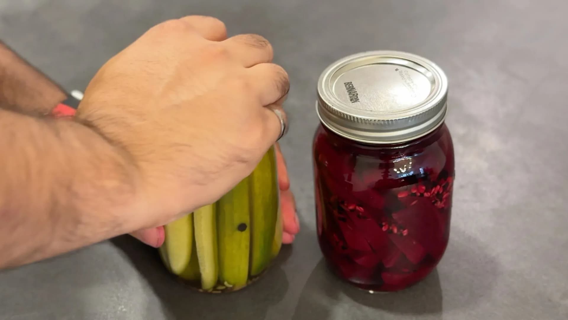 Hand holding fresh green beans next to a jar of deep red pickled vegetables in pickling solution.