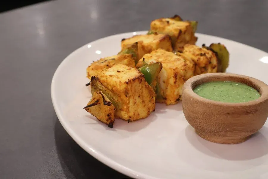 Air fryer paneer tikka skewers on white plate with green cilantro chutney in wooden bowl.
