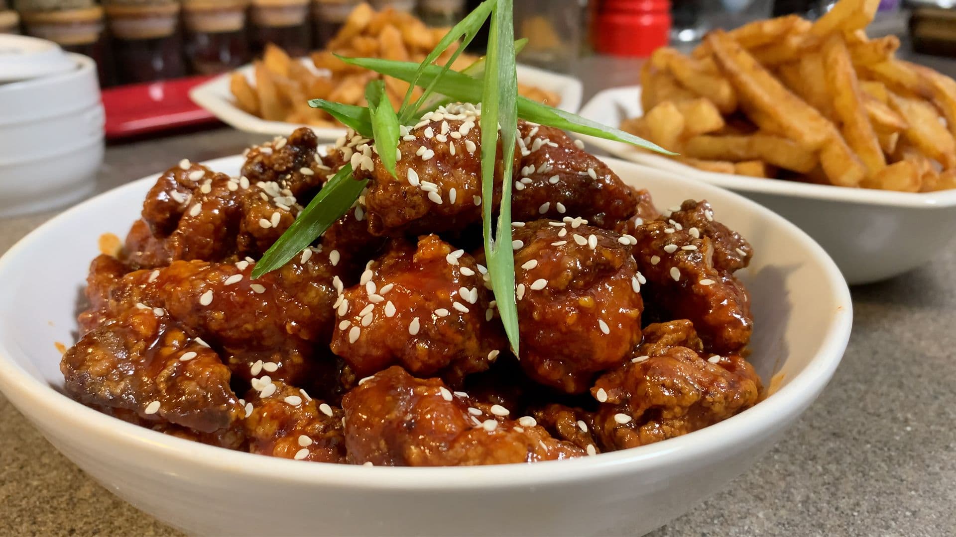 Crispy Korean popcorn chicken coated in sesame seeds and green onions in a white bowl, with fries in the background.