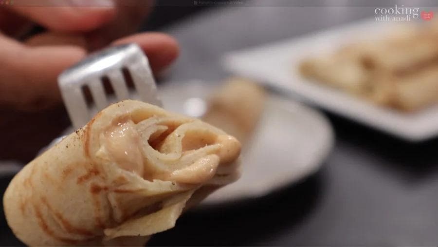 A rolled pumpkin crepe held on a fork, showing its spiral texture and golden-brown color on a plate.