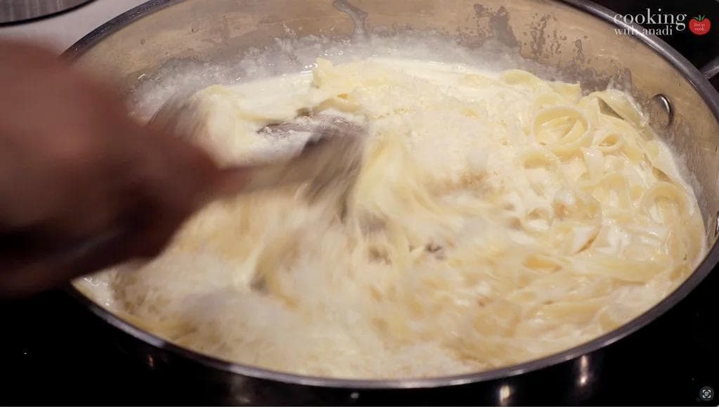Creamy Alfredo sauce being stirred in a pan with melted cheese and pasta, thickening to coat consistency.