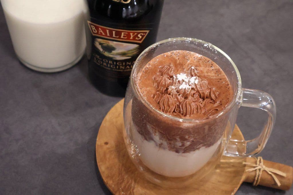 Rich whipped Baileys hot chocolate in a glass mug with cocoa powder on top, served with a Baileys bottle and candle.