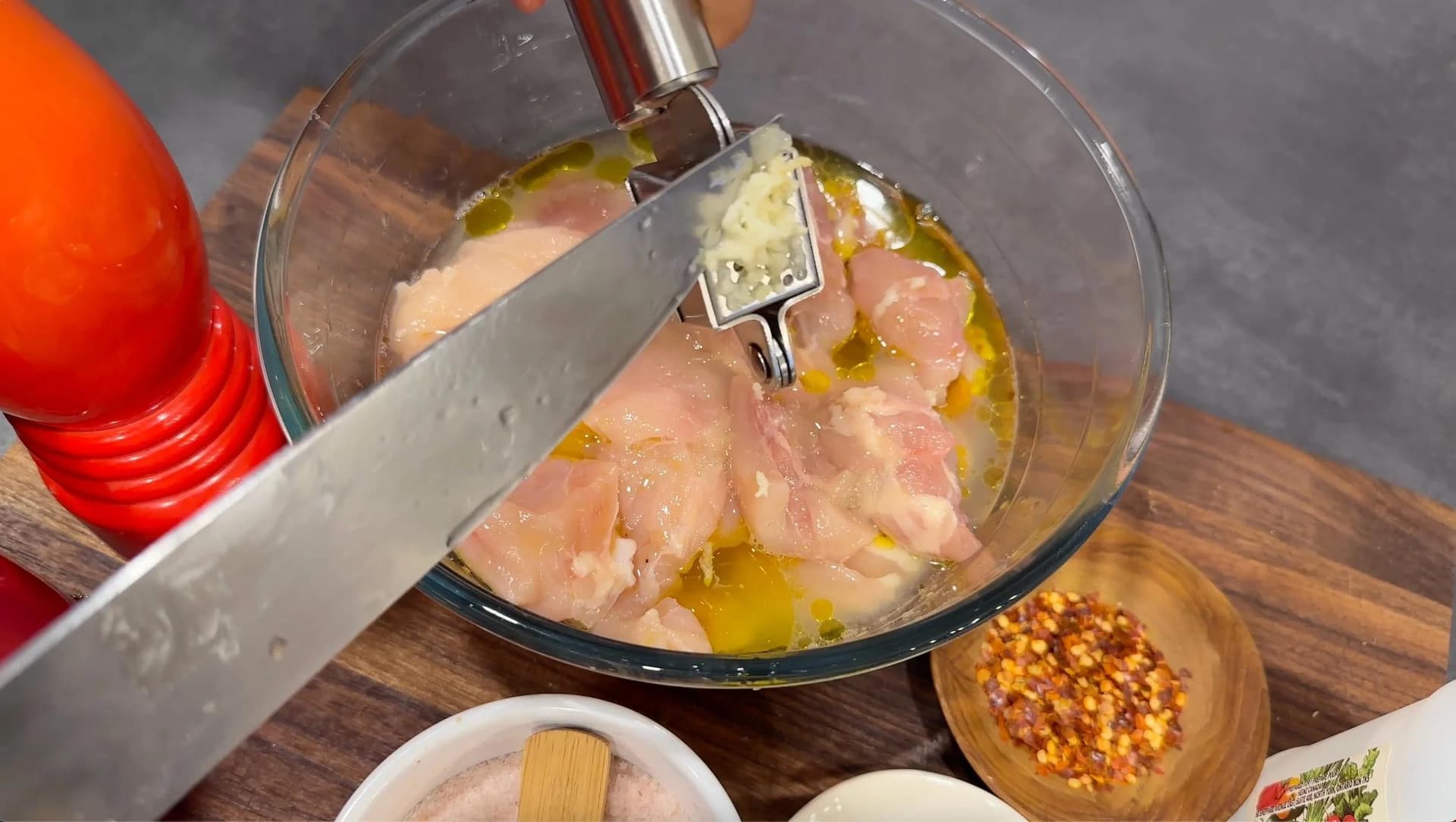 Cubed chicken marinating in olive oil and lemon juice with herbs and spices in a glass bowl, being stirred with a knife.