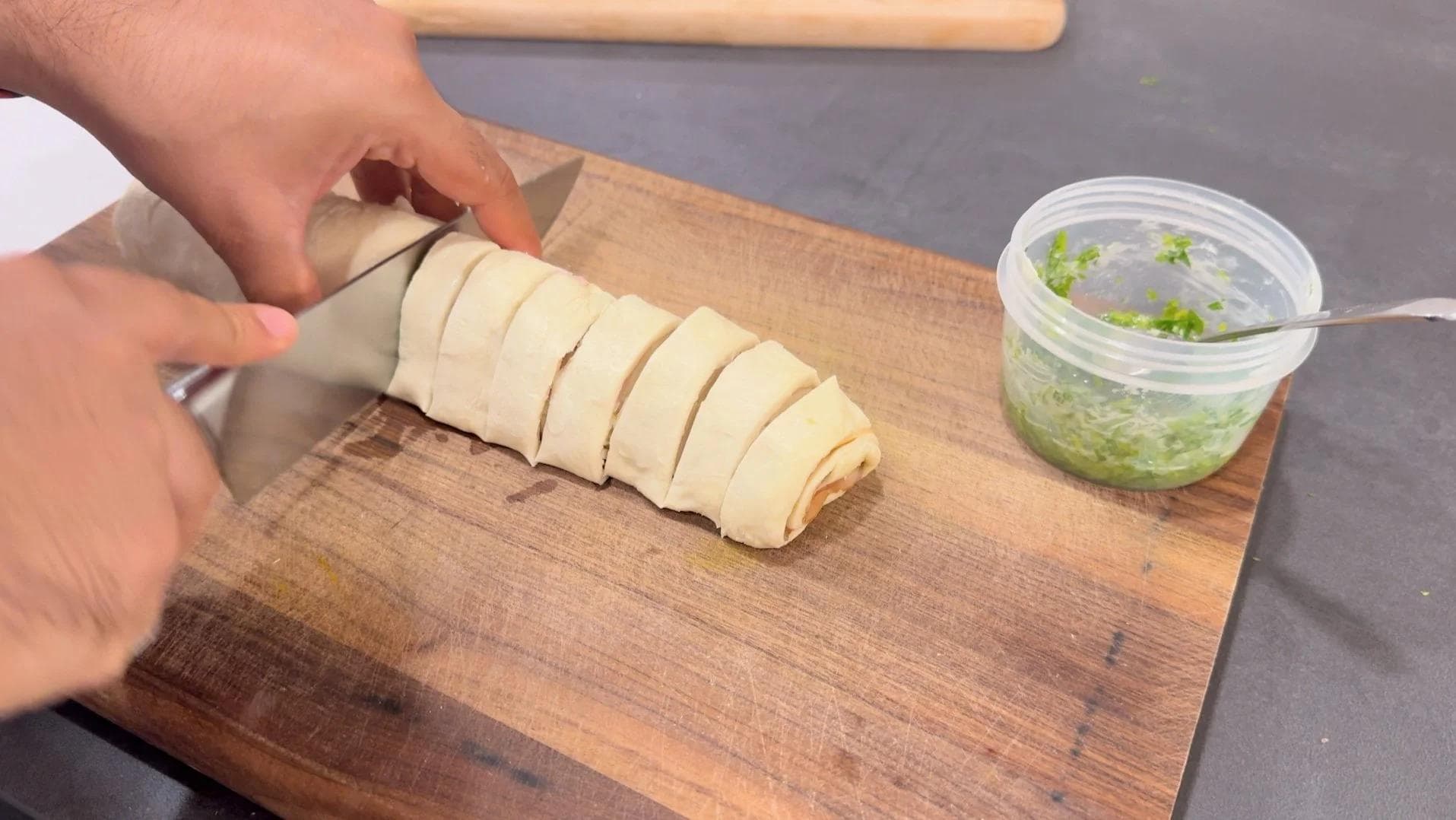 Sliced ham and cheese pinwheel pastry being cut on a wooden board with herb butter in a container nearby.