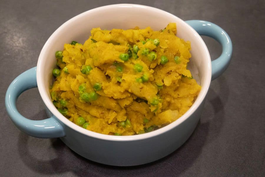 Golden turmeric-spiced potato and pea curry served in a blue-handled white bowl, garnished with fresh green peas.