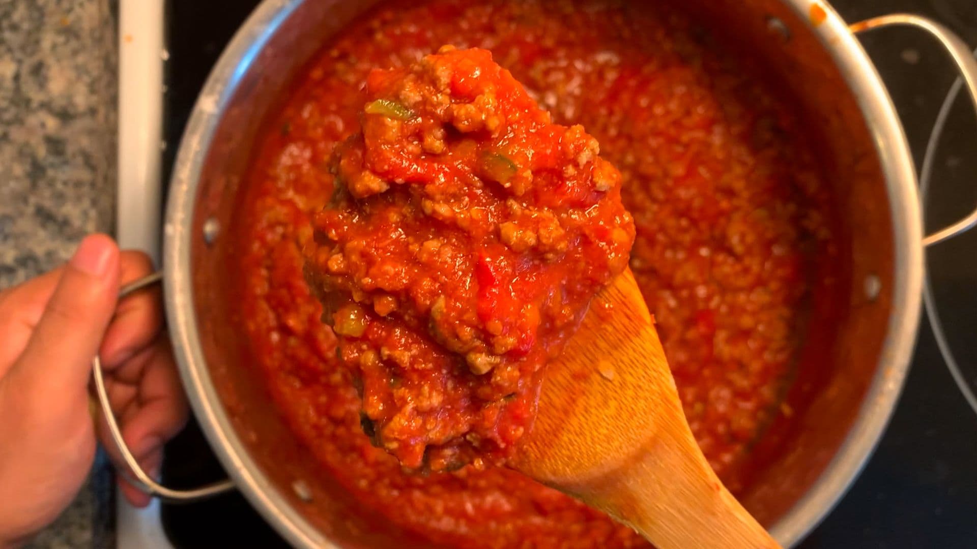 Wooden spoon stirring rich, chunky bolognese sauce in a stainless steel pot on the stove.