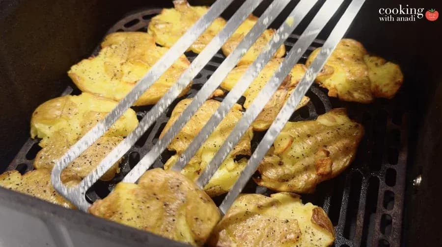 Smashed potatoes cooking on air fryer grates with tongs, golden and seasoned with black pepper.
