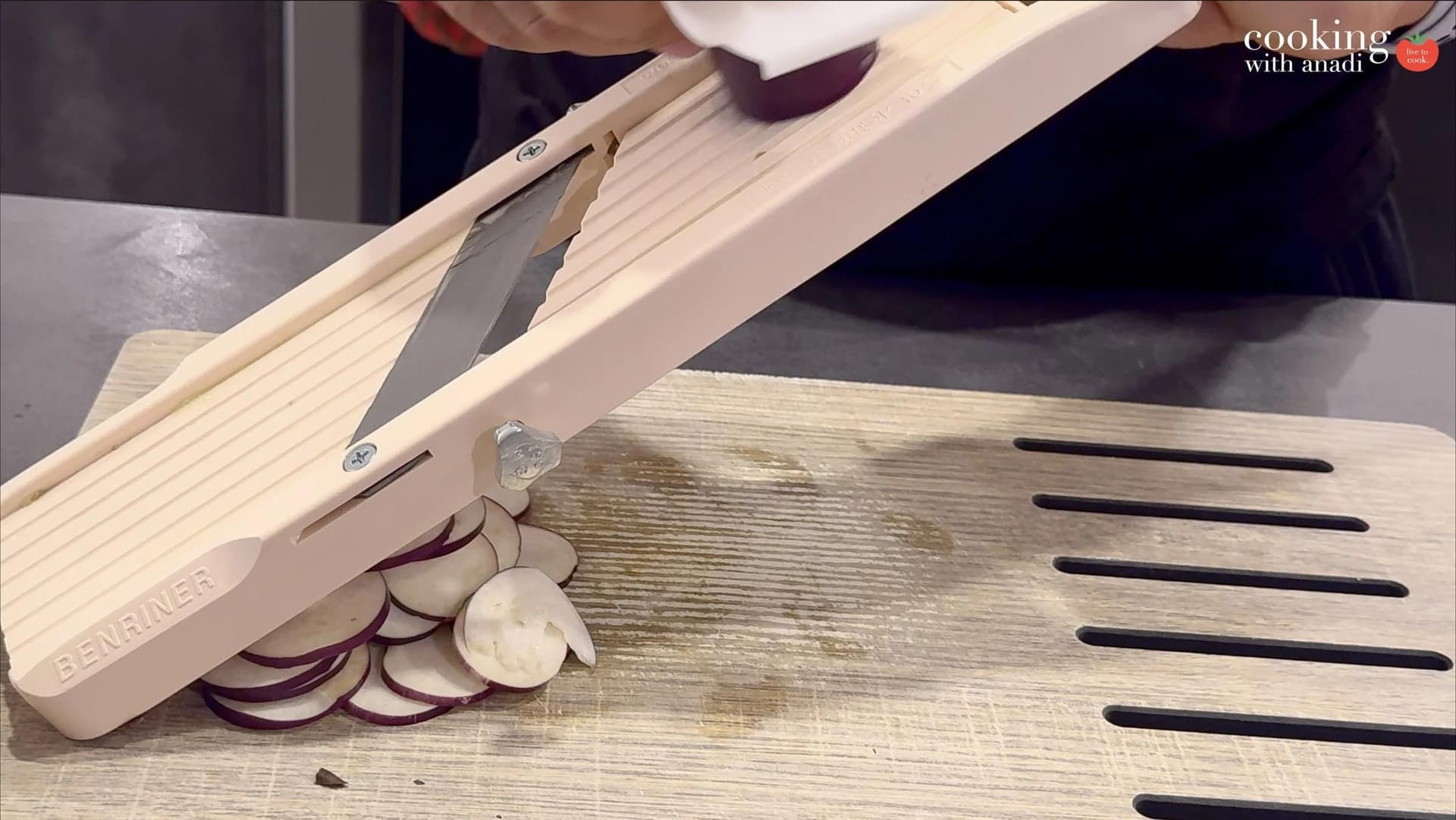 Red onion being sliced into thin rounds on a Benriner mandoline slicer over a wooden cutting board.
