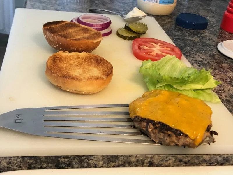 Assembled cheeseburger with melted yellow cheese on a sesame seed bun, served with lettuce, tomato, pickles, and red onion slices on white plate.