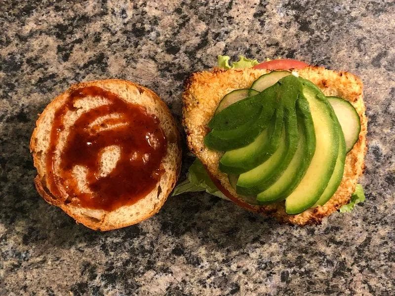 Toasted burger bun halves topped with ketchup on left and sliced avocado on right, on granite countertop.