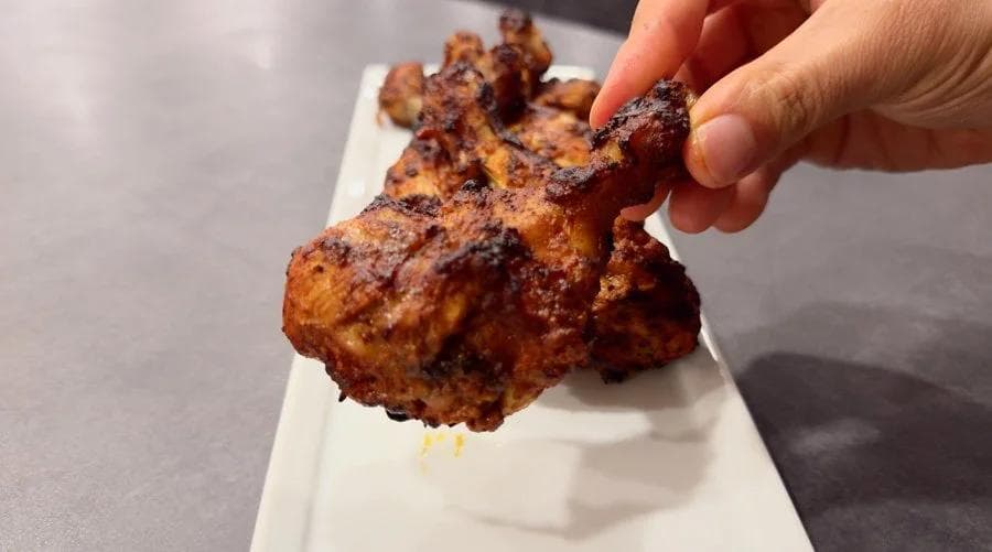 Hand holding a golden-brown air fryer tandoori chicken piece with charred spices on a white plate.