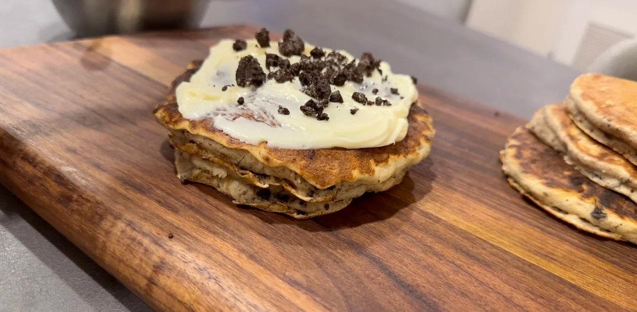 Stacked gluten-free pancakes topped with cream cheese glaze and crushed Oreos on a wooden board.