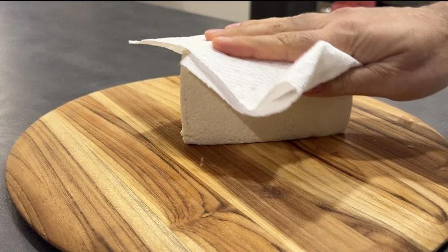 Hand patting tofu block dry with paper towel on wooden cutting board.