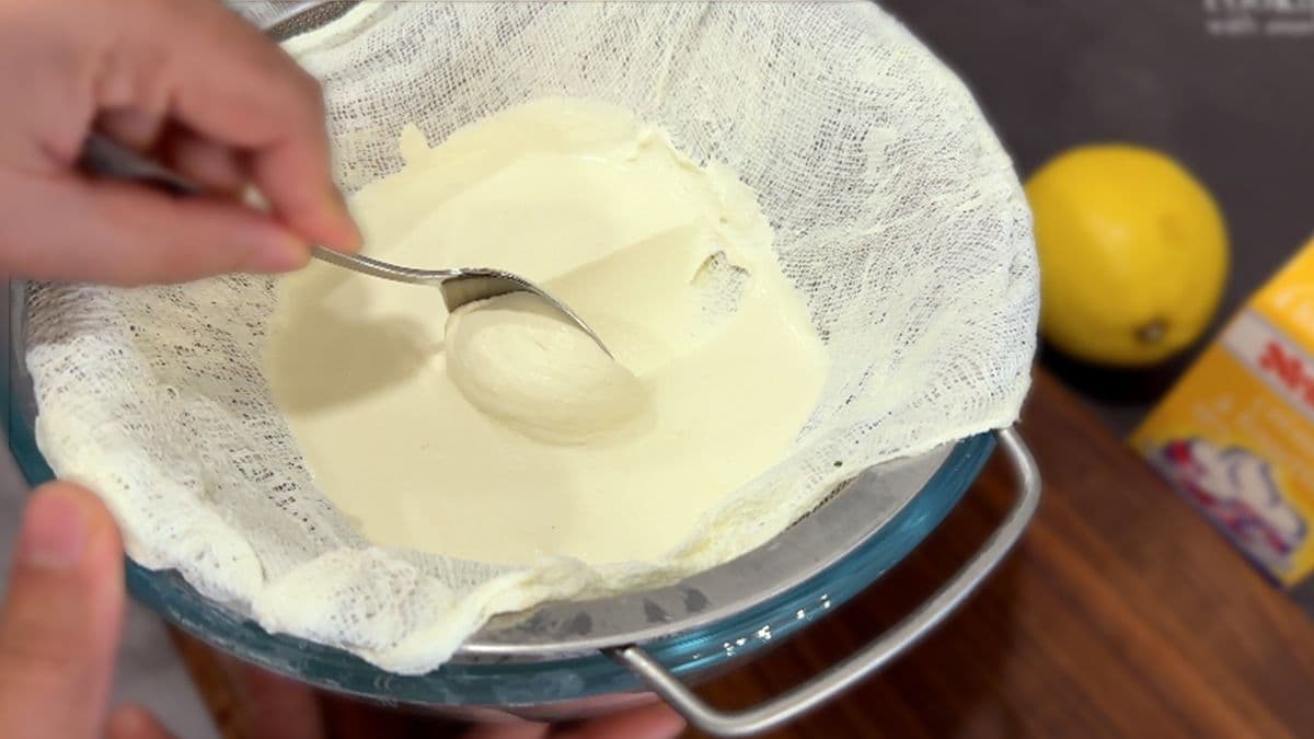 Homemade mascarpone cheese being strained through cheesecloth with a spoon, showing creamy white cheese curds.