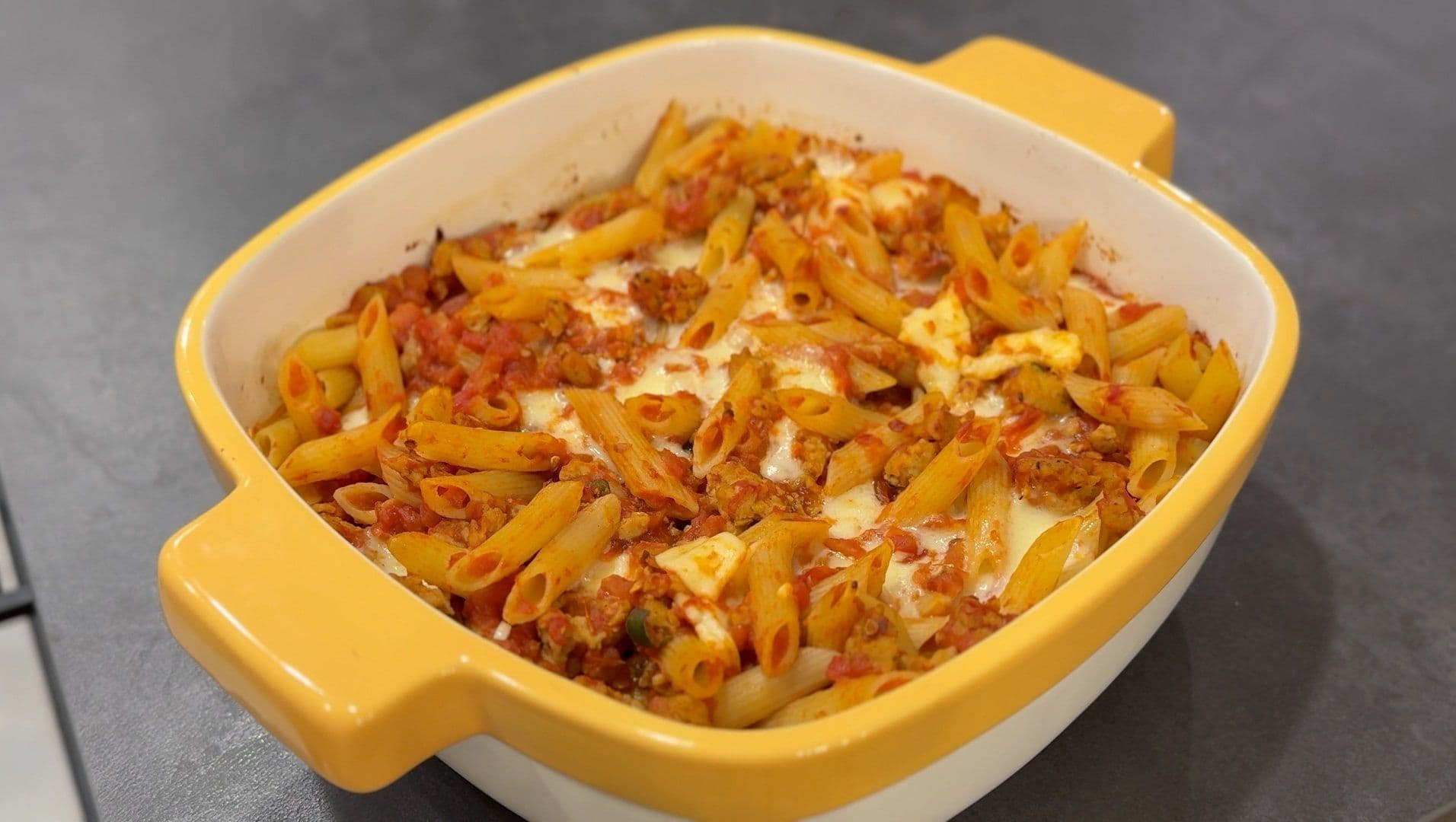 Cheesy baked penne with spicy Italian sausage in a yellow casserole dish with melted mozzarella and provolone.