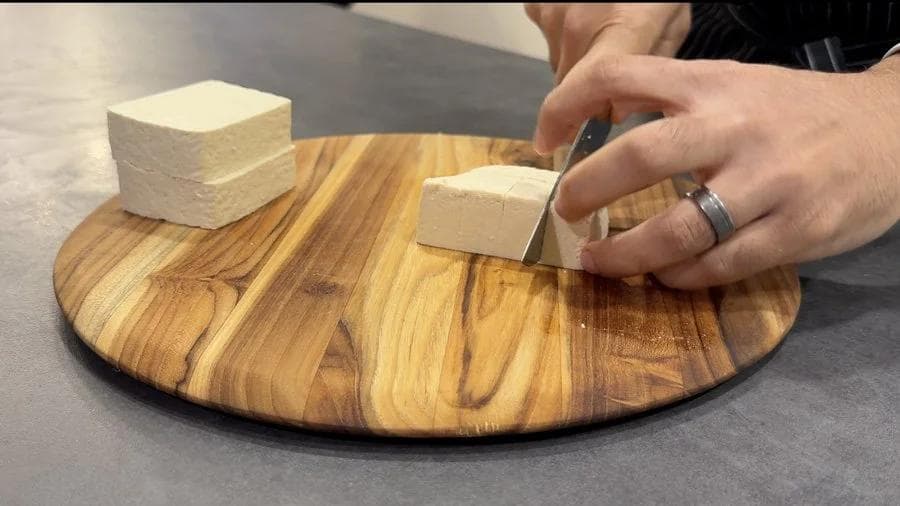 Hands cutting pressed tofu into cubes on a wooden cutting board.