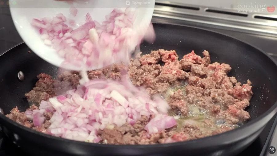 Ground beef being cooked in a skillet with diced pink onions being added from a bowl.