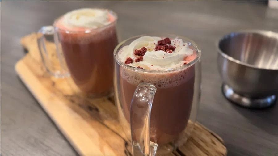 Two mugs of red velvet latte topped with whipped cream and crushed cookies on a wooden board.