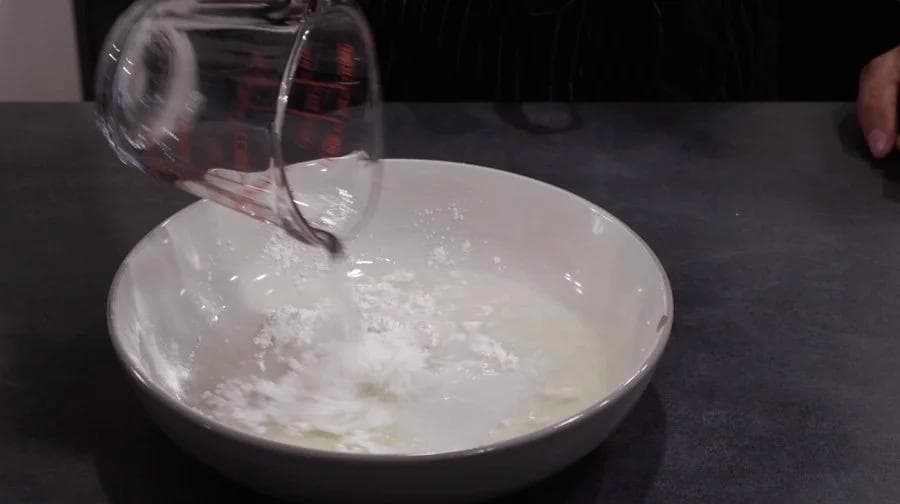 Pouring liquid into a white bowl containing dry breadcrumb mixture for chicken nugget breading preparation.