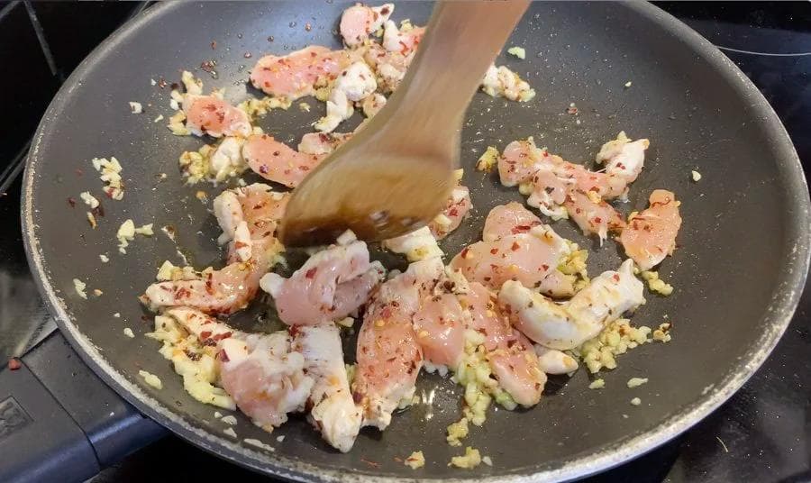 Wooden spoon stirring diced chicken and garlic in a nonstick pan during initial cooking stage.