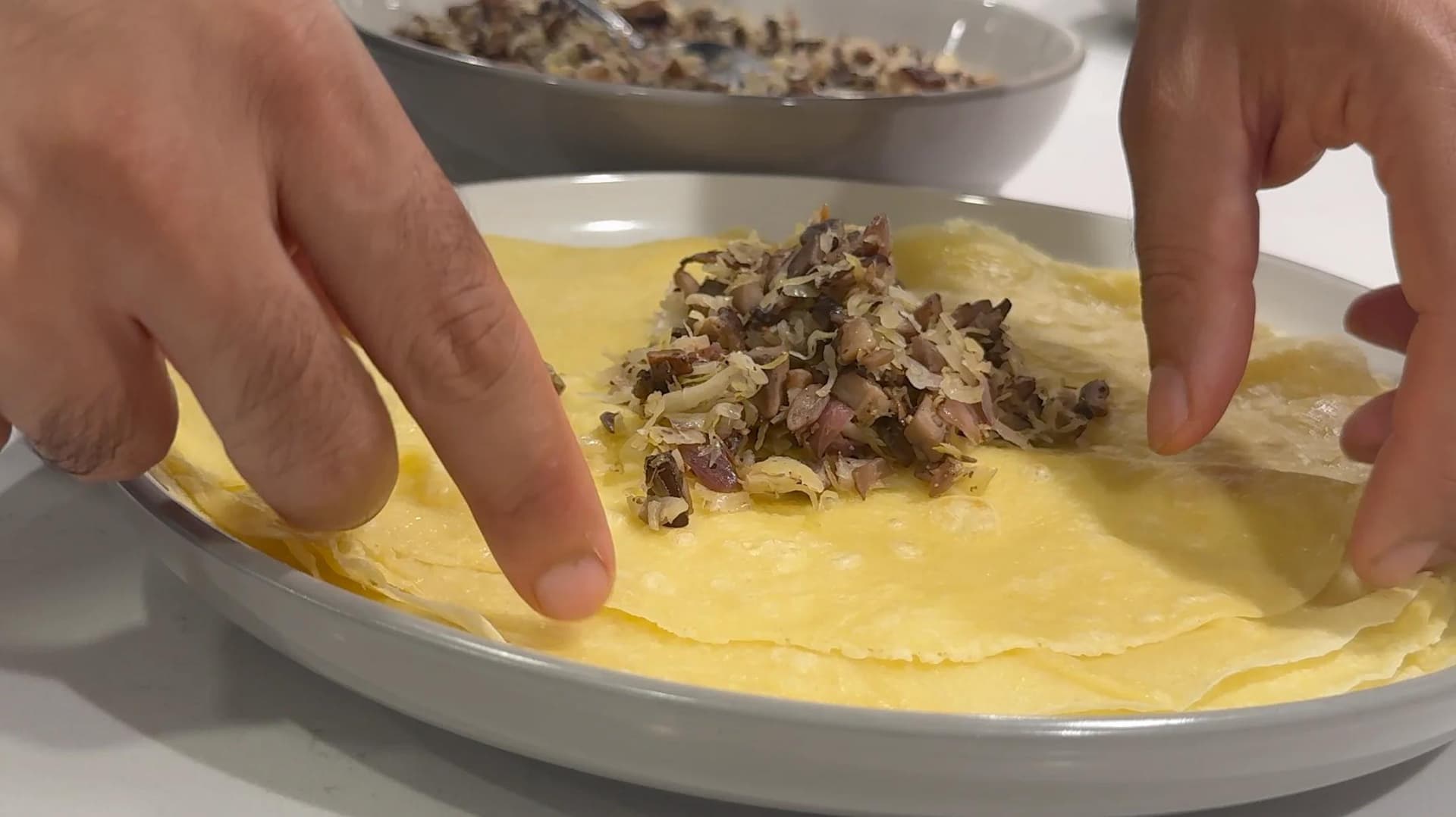Hands folding a crepe with mushroom-sauerkraut filling on a white plate during krokiety preparation.