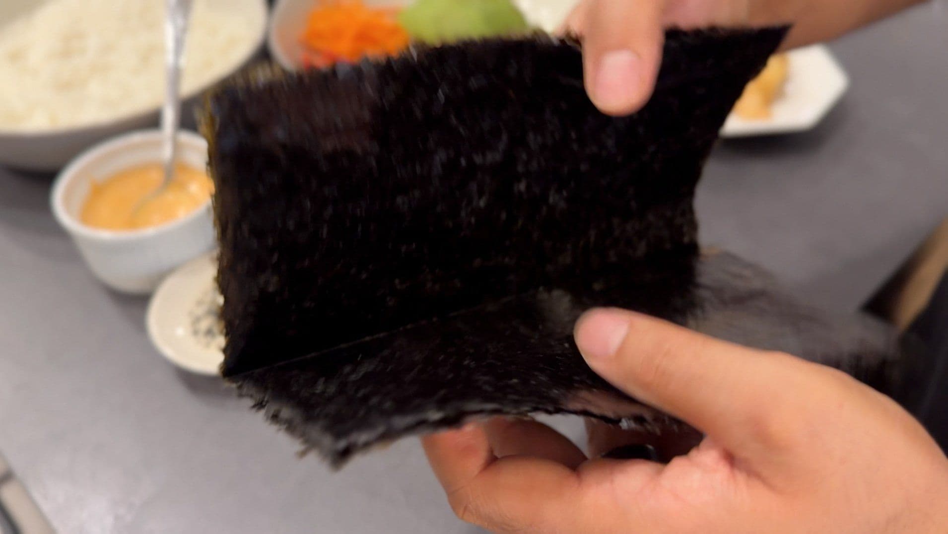 Hand holding a sheet of nori (seaweed) with sushi ingredients and bowls visible in background.