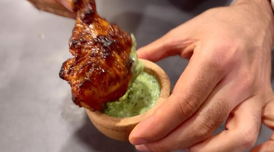 Tandoori chicken being dipped into green mint chutney, held in a hand over a small bowl.
