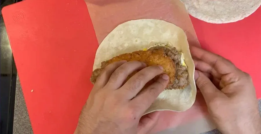 Hands wrapping a breakfast wrap filled with egg patties and hashbrown in parchment paper on a red surface.