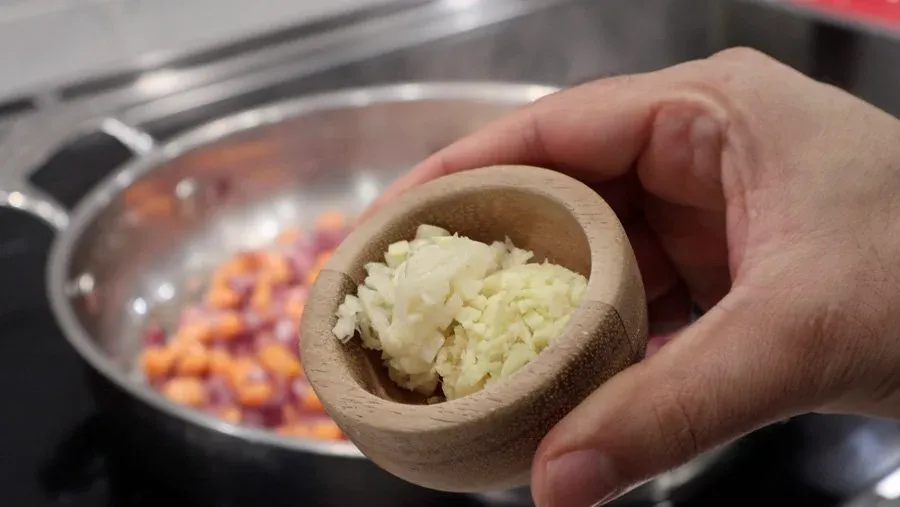 Hand holding wooden mortar with minced garlic over pot of sautéed onions and carrots on stovetop.