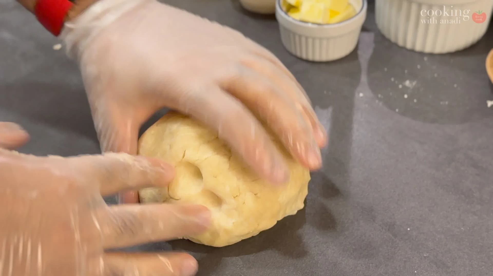 Hands kneading yellow pie dough on a dark surface with ramekins of filling in background.