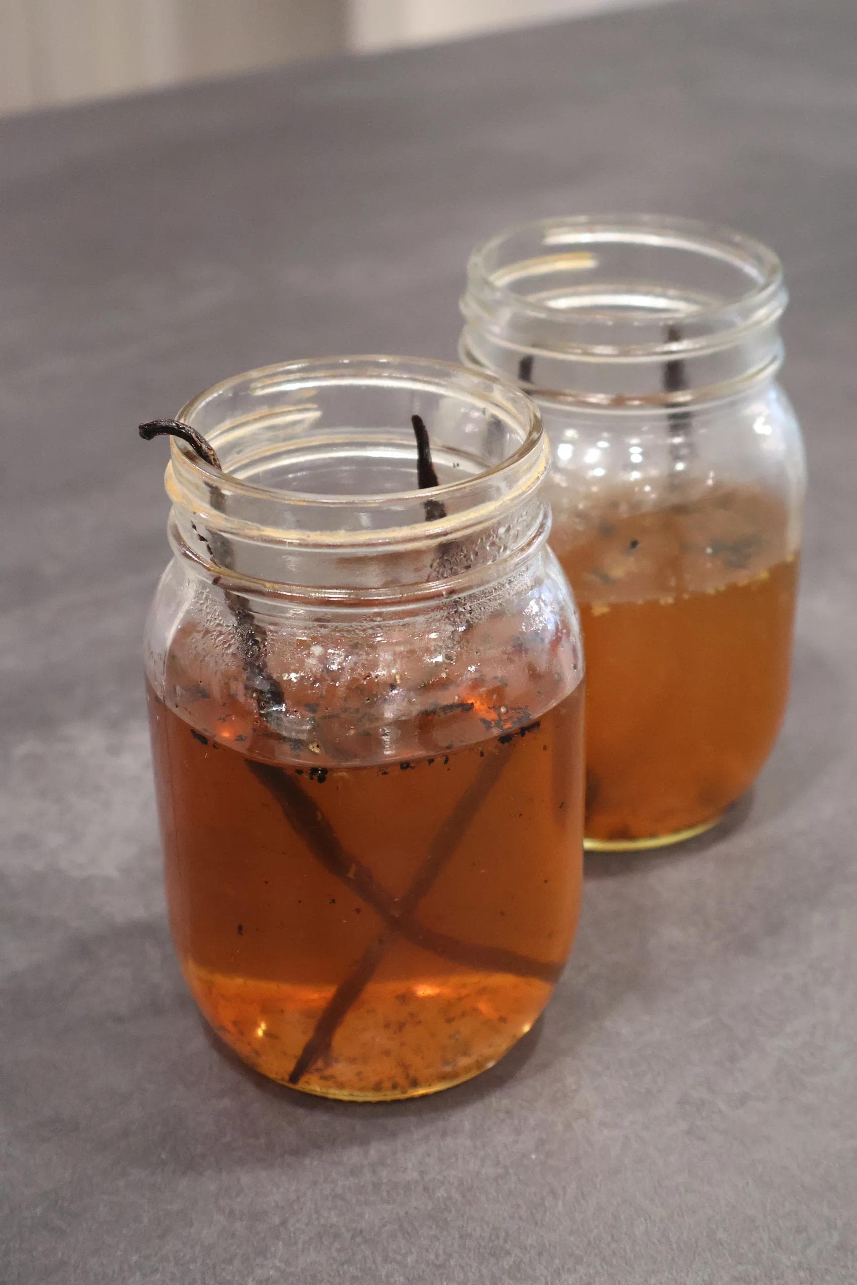 Two mason jars with vanilla syrup and vanilla beans, one with ice and a straw, sitting on a gray surface.