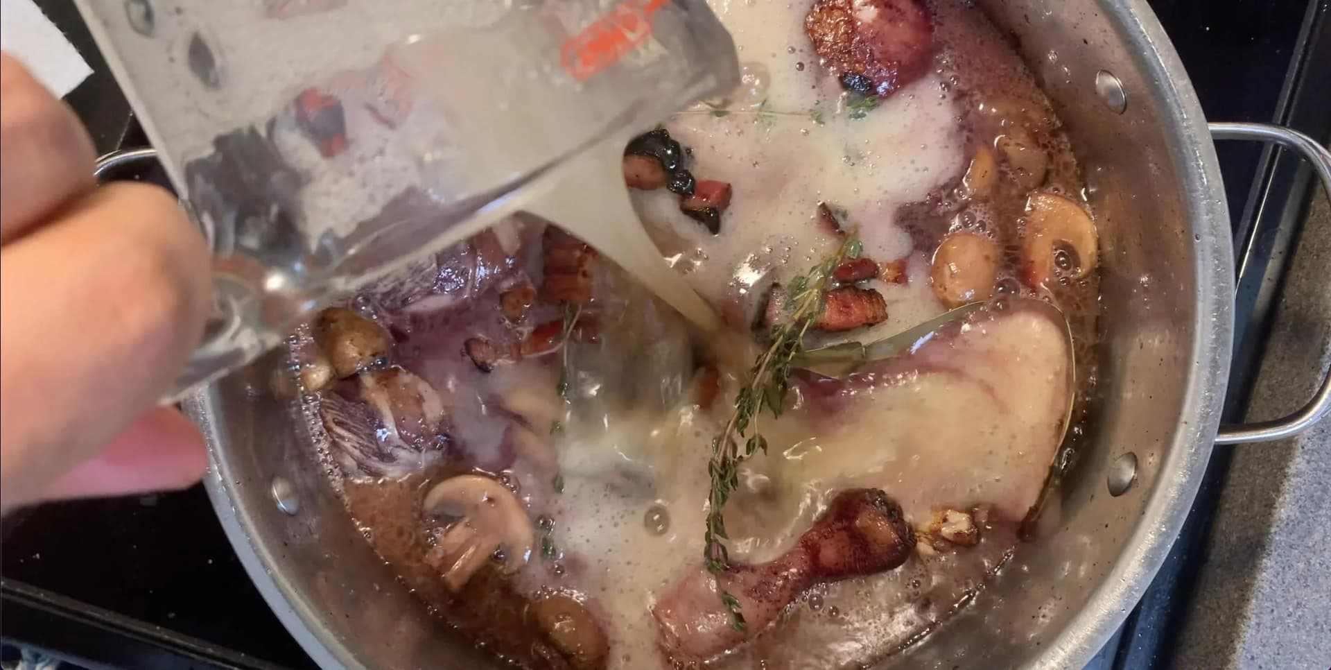 Liquid being poured into a pot of browned chicken, mushrooms, bacon, and herbs for coq au vin.