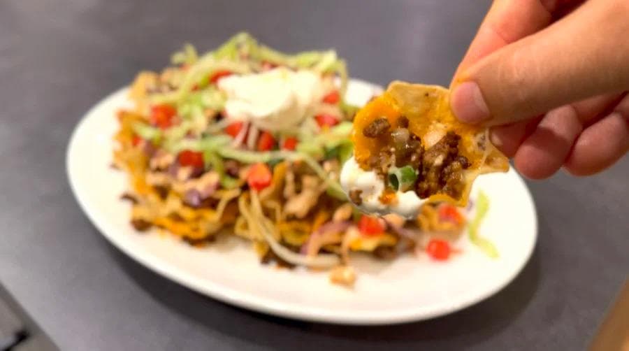 Hand holding a crispy nacho chip topped with melted cheese, burger meat, sour cream, and fresh toppings over a plate of cheeseburger nachos.