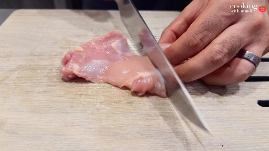 Hands cutting raw chicken thigh into bite-sized pieces on a wooden cutting board with a chef's knife.