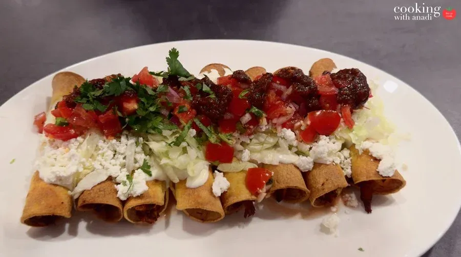 Crispy taquitos on a white plate topped with crema, shredded lettuce, cotija cheese, pico de gallo, salsa roja, and cilantro.
