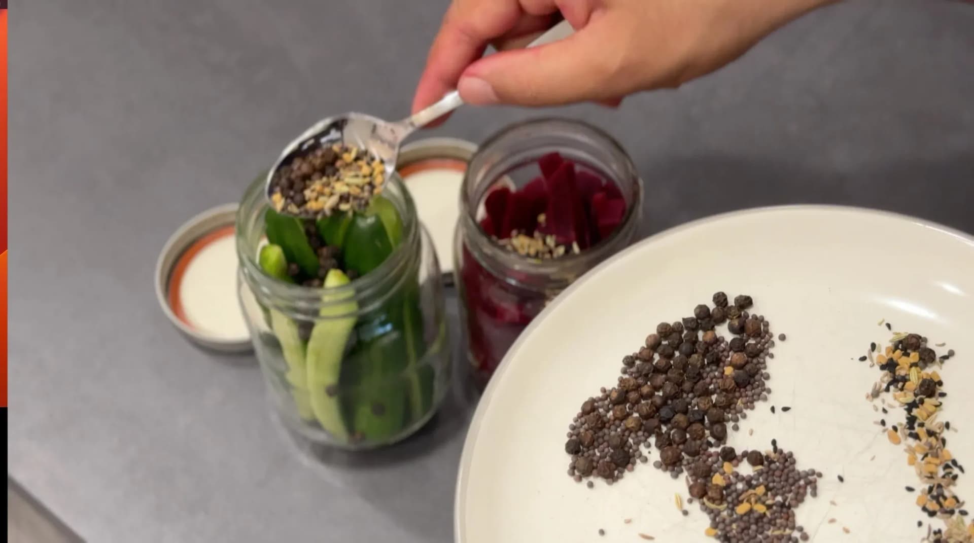 Hand spooning whole spices into mason jar with blanched green beans and other vegetables.