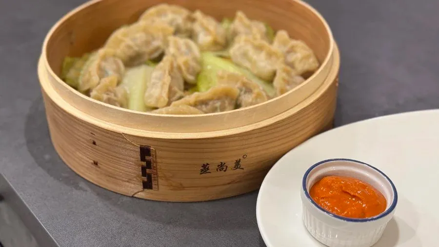 Steamed chicken momos in a bamboo steamer basket with a small bowl of orange dipping sauce on the side.