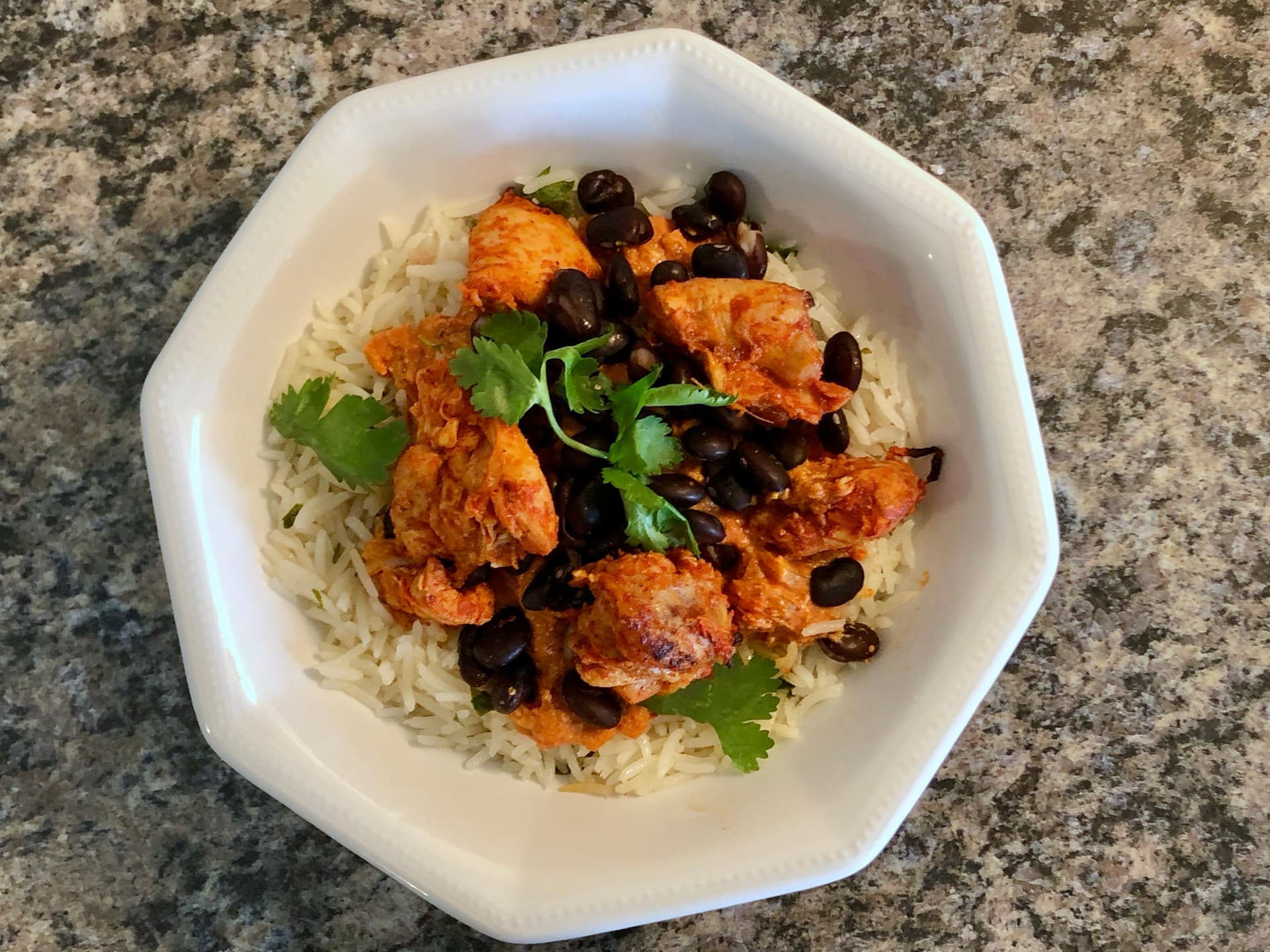 Chicken tikka masala bowl with rice, black beans, spiced chicken pieces, and cilantro garnish on white plate.